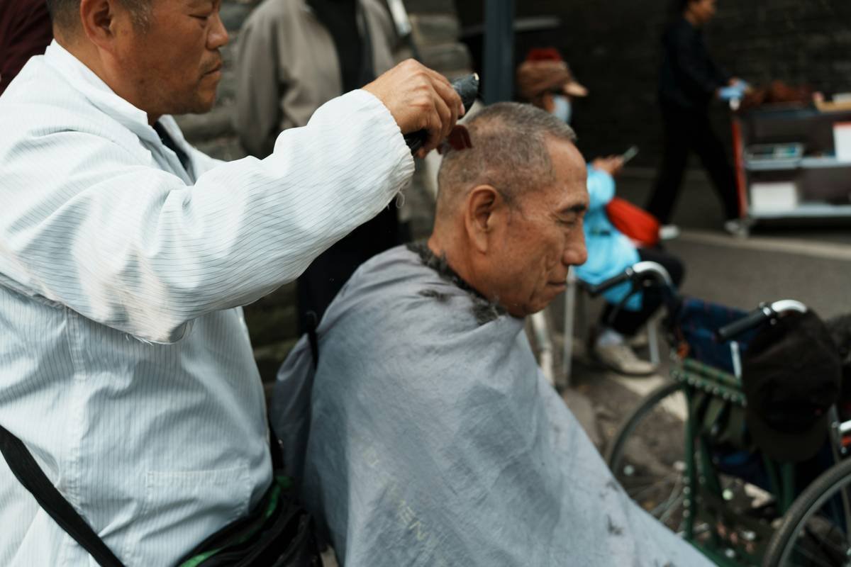 Man getting a haircut outdoors from another man.